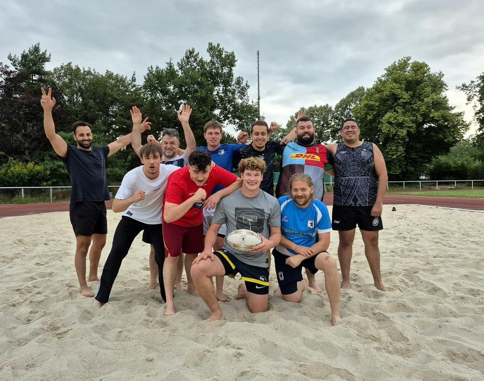 Gruppenshot von zehn Rugbyspielern auf einem Sandfeld, einige posieren, während andere jubeln.
