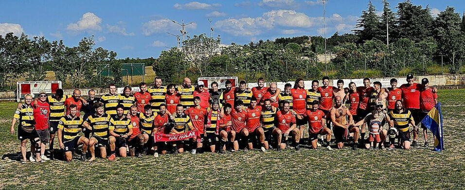 Gruppenfoto von Rugby-Spielern in roten und schwarzen Trikots sowie gelben und schwarzen Streifen, auf einem grünen Spielfeld.