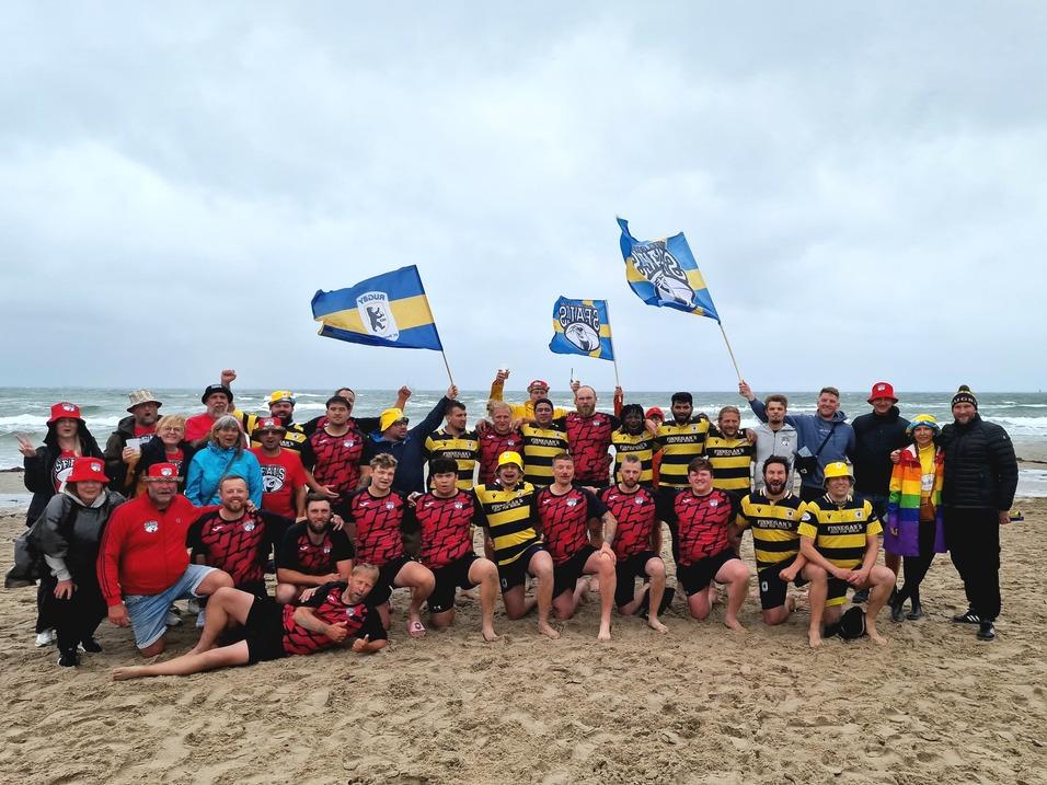 Gruppenshot von Rugbyspielern und Fans am Strand, die Flaggen schwenken und gemeinsam feiern.