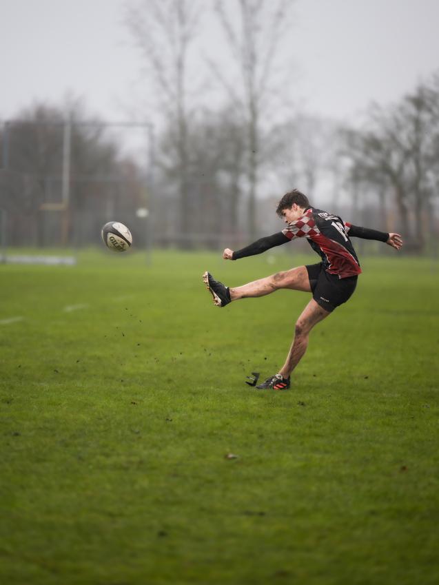 Ein Rugbyspieler tritt einen Ball auf einem nassen, grünen Spielfeld, während er in Aktion ist.