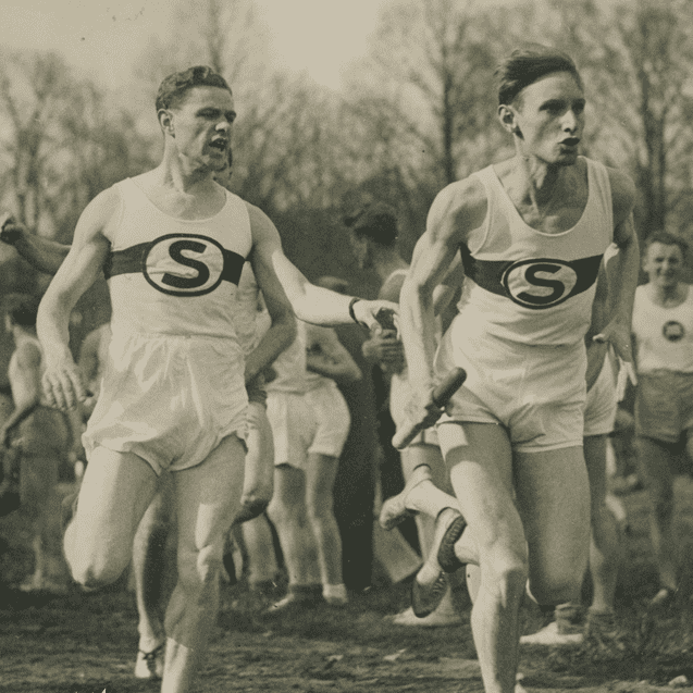 Zwei Läufer in historischen Trikots übergeben einen Staffelstab bei einem Wettkampf auf einem staubigen Sportplatz.