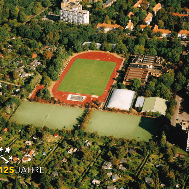 Luftaufnahme eines Sportgeländes mit Fußballfeld und angrenzenden Wohngebieten, umgeben von Bäumen.