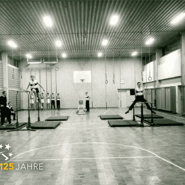 Sporthalle mit Sportlern, die an Turngeräten trainieren, Zuschauer sitzen auf Bänken im Hintergrund. Schwarz-Weiß-Foto.