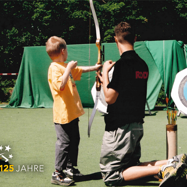 Ein Kind mit Bogen wird von einem Trainer zur richtigen Haltung angeleitet, im Hintergrund eine Zielscheibe.