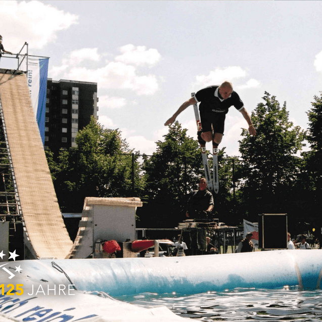 Mann springt mit Stelzen von einer Rampe in ein Wasserbecken, Zuschauer verfolgen das Geschehen im Hintergrund.
