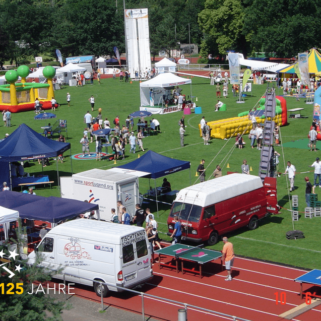 Blick auf eine Veranstaltung im Freien mit mehreren Ständen, Spielen und Menschen auf einer grünen Rasenfläche.