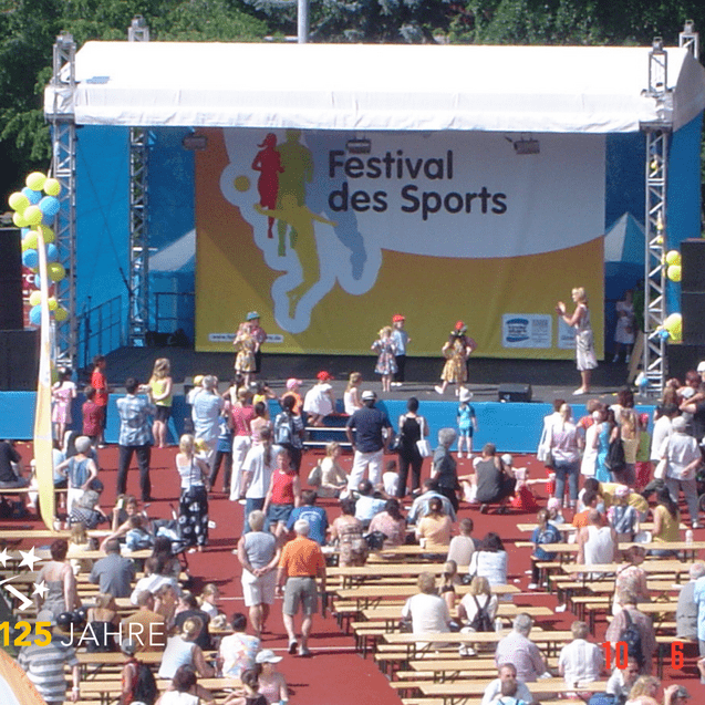 Menschenmenge bei einem Sportfestival, mit einer Bühne und Tänzern in bunten Kostümen unter Sonnenschein.