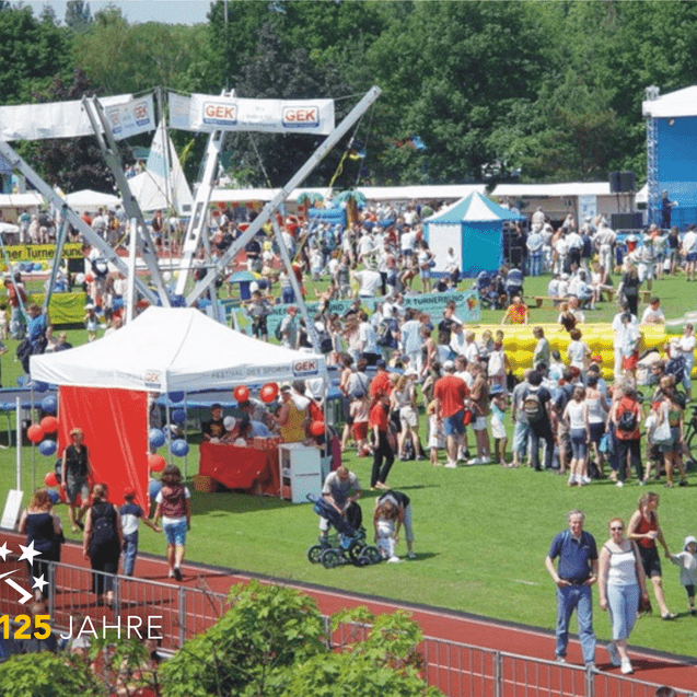 Voller Veranstaltungsgelände mit Menschen, Zelten, Attraktionen und einer Bühne im Hintergrund auf einer Sportwiese.