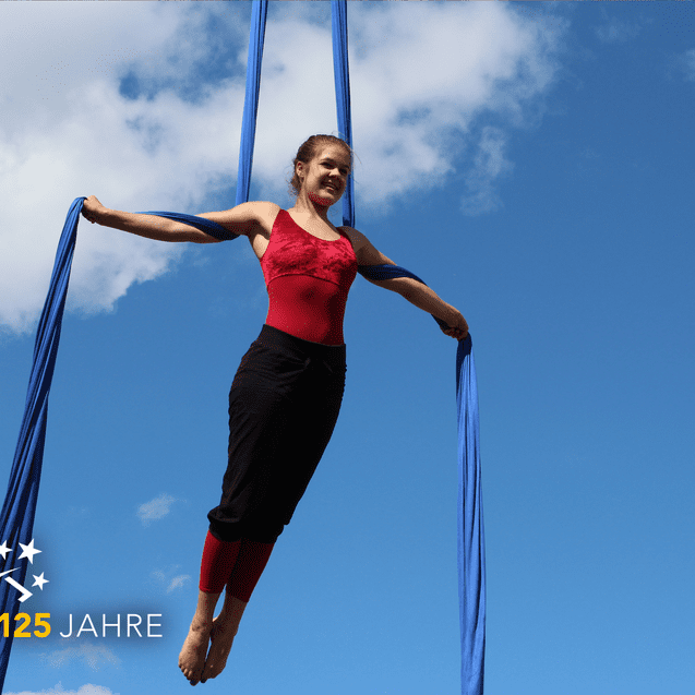 Frau in rotem Oberteil schwebt an blauen Tüchern vor blauem Himmel mit Wolken. 125 Jahre Jubiläumslogo unten links.