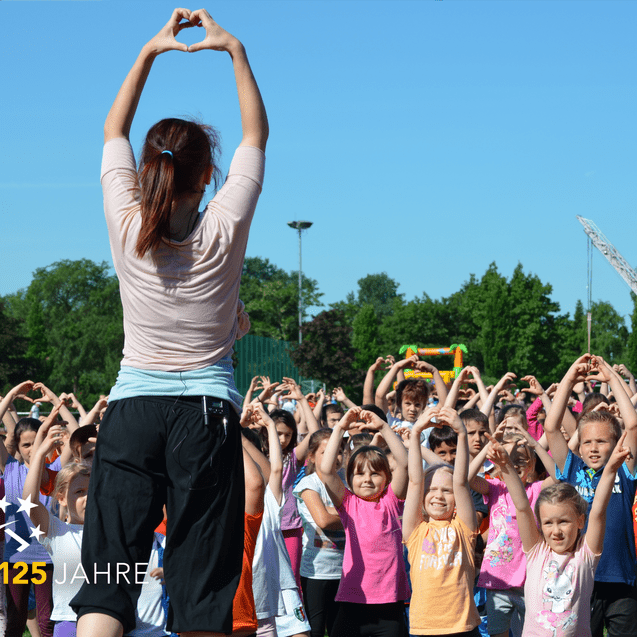Gruppentanz auf einem Feld mit Kindern, die Herzen mit den Händen formen, angeleitet von einer Trainerin.