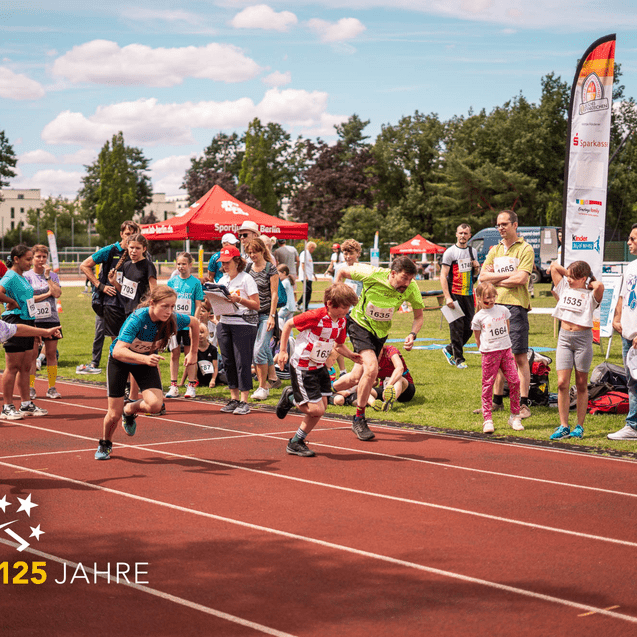 Kinder und Jugendliche starten beim Wettlauf auf einer roten Laufbahn, Publikum und Betreuer beobachten den Wettbewerb.