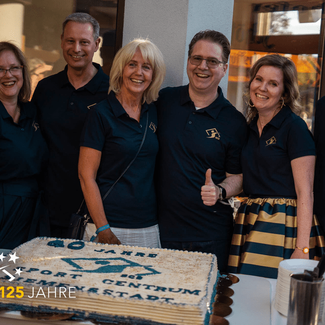 Sechs Personen in schwarzen Polo-Shirts stehen lächelnd neben einer großen Geburtstagstorte mit der Aufschrift „125 Jahre“.