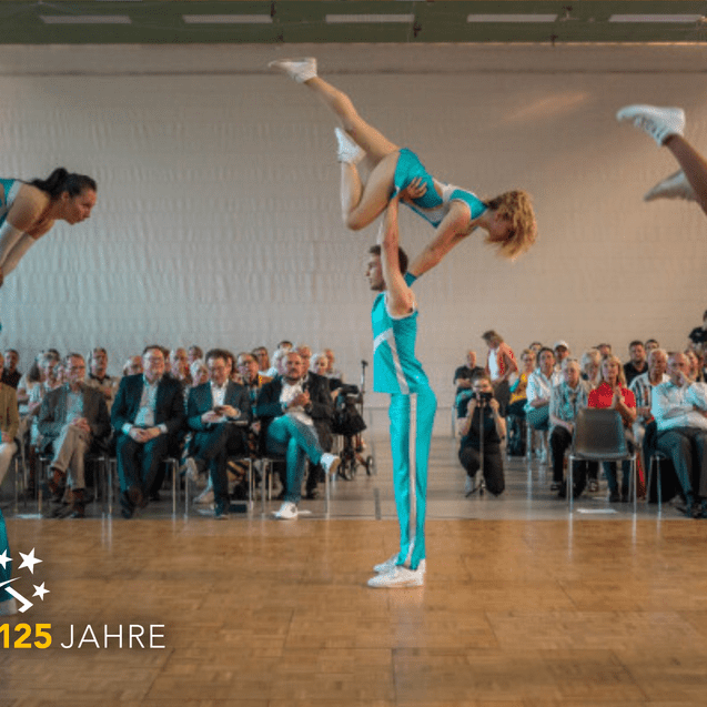 Vier akrobatische Tänzer in türkisfarbenen Outfits führen Hebefiguren in einer Aufführung vor Publikum auf.