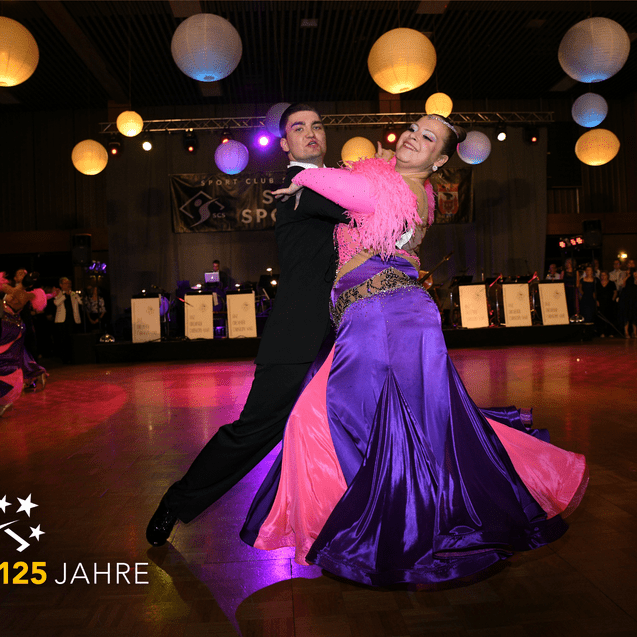 Tanzpaar in eleganten Kostümen tanzt auf einer festlichen Veranstaltung mit bunten Laternen im Hintergrund.