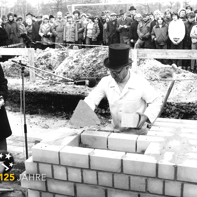 Ansprache eines Mannes am Mikrofon, während ein anderer Bauarbeiter Mauersteine auf einer Baustelle setzt. Zuschauergruppe im Hintergrund.