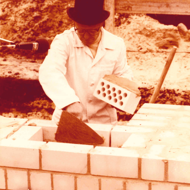 Mann in Zylinder und weißem Laborkittel verlegt einen Baustein auf einer Mauer, umgeben von Zuschauern.