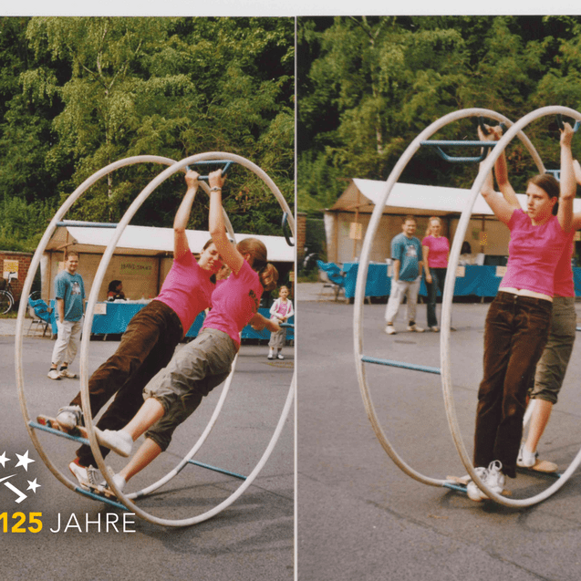 Zwei Personen in pinken T-Shirts balancieren in einem großen Spielreifen auf einem Platz mit Ständen im Hintergrund.