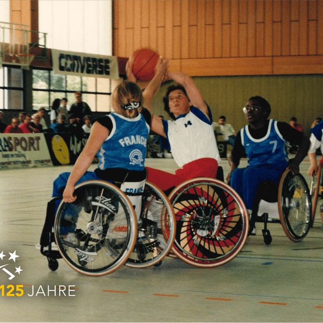 Vier Basketballspieler im Rollstuhl kämpfen um den Ball auf dem Spielfeld während eines Wettkampfs.