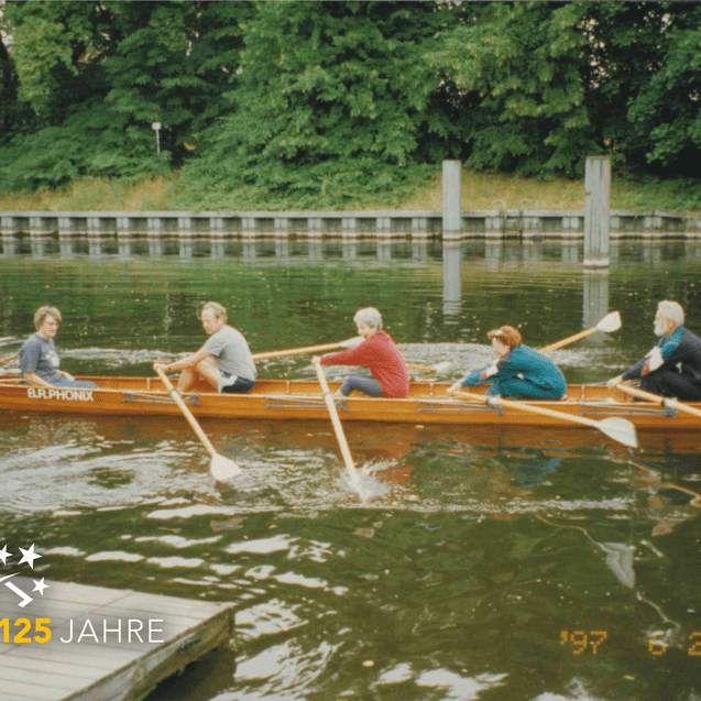 Fünf Ruderer in einem Holzboot auf einem ruhigen Gewässer, umgeben von Bäumen und einem Holzsteg im Hintergrund.