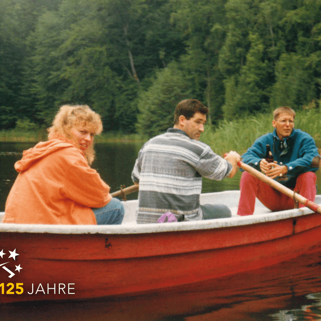 Drei Personen in einem roten Boot auf einem ruhigen Gewässer, umgeben von Waldvegetation.