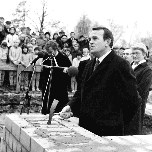 Mann in Anzug spricht vor einer Menschenmenge, während er einen Ziegelstein auf einem Baugrundstück platziert.