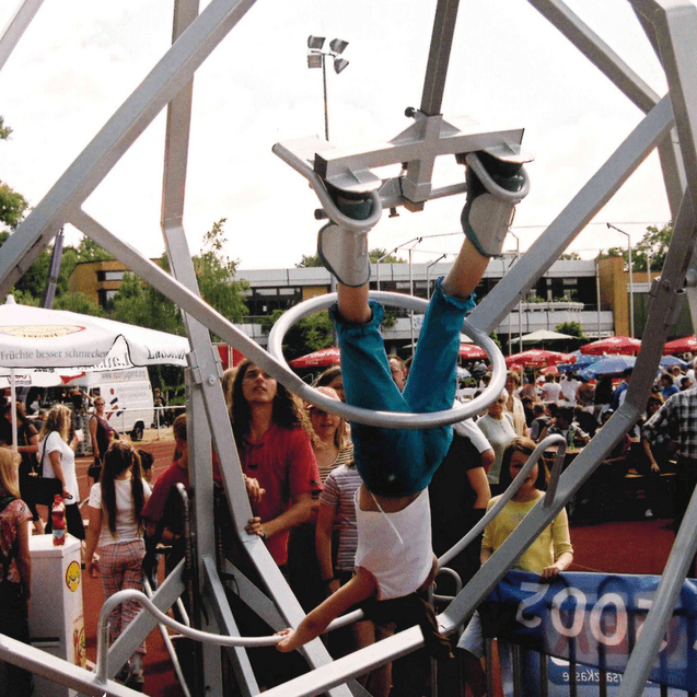 Ein Kind hängt kopfüber in einem Spielgerät, umgeben von Menschenmenge auf einem Fest.