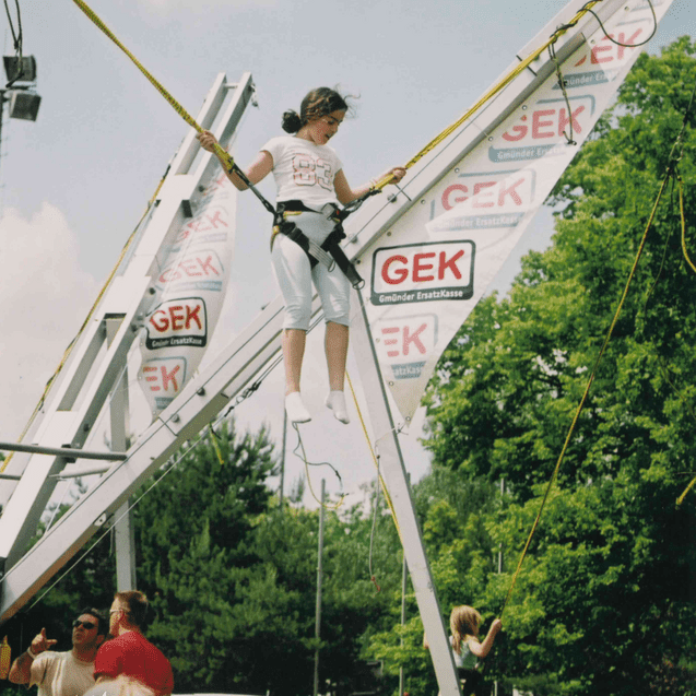 Mädchen in Sicherheitsgurt schwebt an einem Trampolin-Gerüst über dem Boden, umgeben von Zuschauern bei einer Veranstaltung.