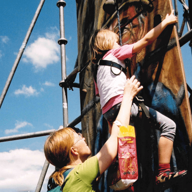 Ein Kind klettert an einem bemalten Kletterturm, während eine Erwachsene hilfsbereit darunter steht.