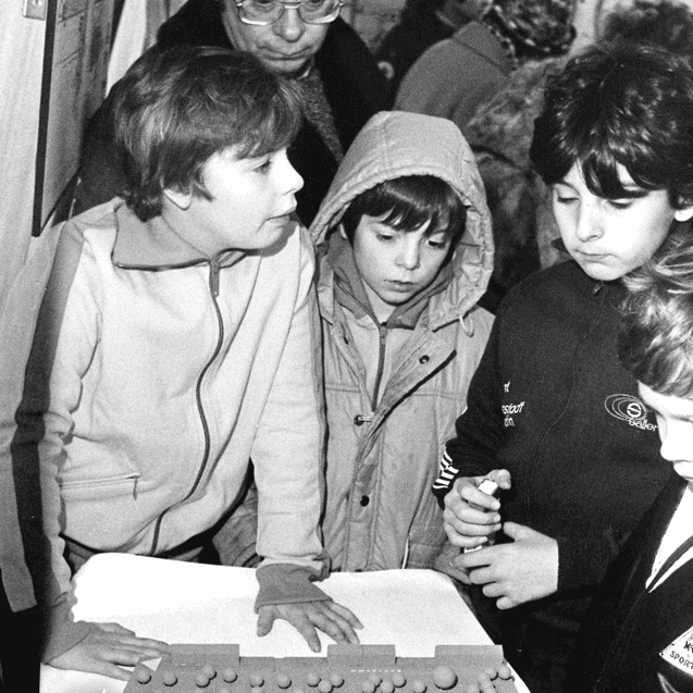 Gruppe von Kindern und Erwachsenen betrachtet ein Modell auf einem Tisch, umgeben von Zeltwänden.