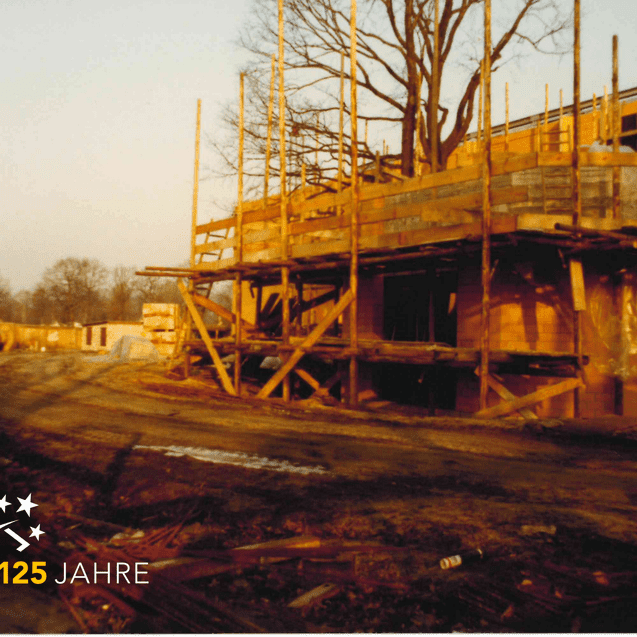 Baustelle mit Holzgerüst und unvollendeter Wandstruktur unter einem Baum im Abendlicht. Mauersteine liegen vor dem Gebäude.