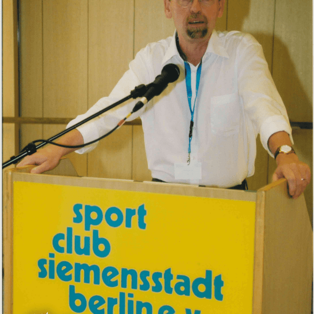 Mann mit Brille hält eine Rede am Mikrofon vor einem Podium mit der Aufschrift "sport club siemensstadt berlin e.V."