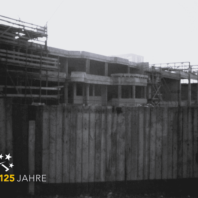 Baustelle eines Gebäudes mit Gerüst und Holzschutzwand, versehen mit einem Logo zum 125-jährigen Jubiläum.