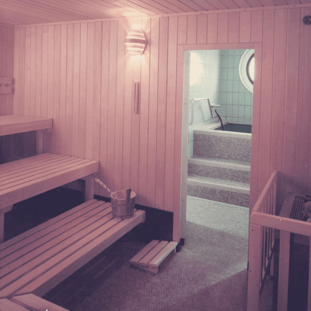 Innenansicht einer Sauna mit Holzbank, Eimer und Zugang zu einem Flur mit einem Schwimmbecken.
