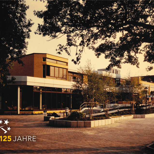 Modernes Geb&auml;ude mit gro&szlig;en Fenstern, umgeben von B&auml;umen und Wegen, im Hintergrund ein Logo mit &bdquo;125 Jahre&ldquo;.