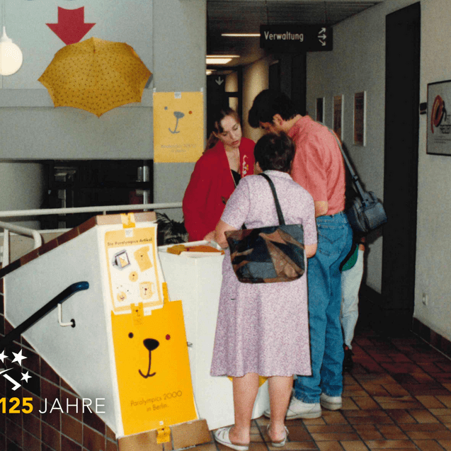 Drei Personen stehen an einem Informationsstand mit einem gelben Plakat für die Paralympics 2000 in Berlin.