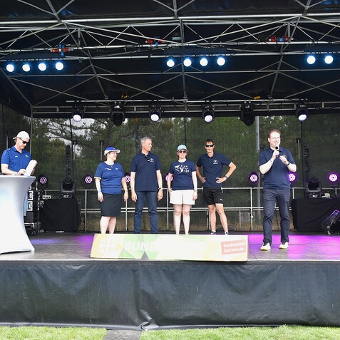 F&uuml;nf Personen stehen auf einer B&uuml;hne im Freien, w&auml;hrend einer von ihnen spricht. Scheinwerfer beleuchten das Event.
