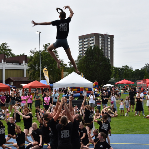 Akrobatin springt von einer Menschengruppe in schwarz gekleideten Cheerleadern w&auml;hrend einer Vorf&uuml;hrung im Freien.