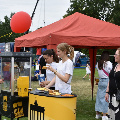 Drei Frauen am Popcornstand, ein Junge greift nach Popcorn, roter Ballon &uuml;ber dem Stand. Im Hintergrund sind Besucher und Spiele.
