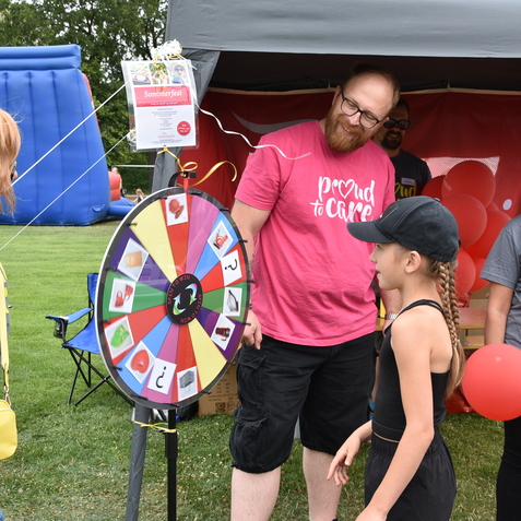 Vier Personen stehen an einem bunten Gl&uuml;cksrad auf einem Sommerfest im Freien, umgeben von roten Luftballons.