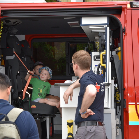 Kind sitzt in einem Feuerwehrfahrzeug, w&auml;hrend ein Feuerwehrmann mit einem Gespr&auml;chspartner spricht.