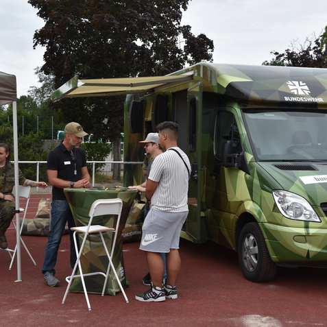 Stand mit Informationsbooth der Bundeswehr, zwei Personen im Gespr&auml;ch, ein Soldat und ein Fahrzeug im Hintergrund.