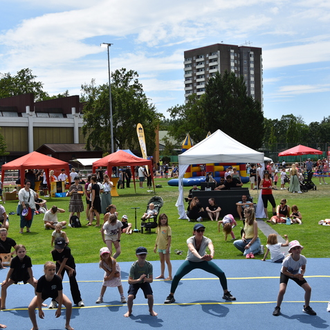 Gruppen von Menschen, darunter Kinder, &uuml;ben aktiv in Sportbekleidung auf einem gr&uuml;nen Rasenplatz w&auml;hrend einer Veranstaltung.