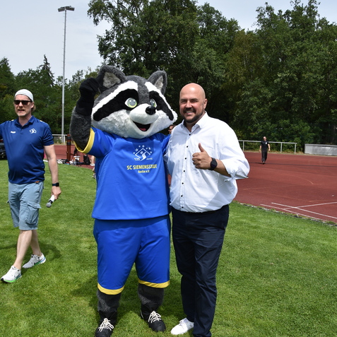 Ein Mann in Wei&szlig; posiert mit einem Waschb&auml;ren-Maskottchen in blauen Sportoutfits auf einer Wiese. Im Hintergrund stehen weitere Personen.