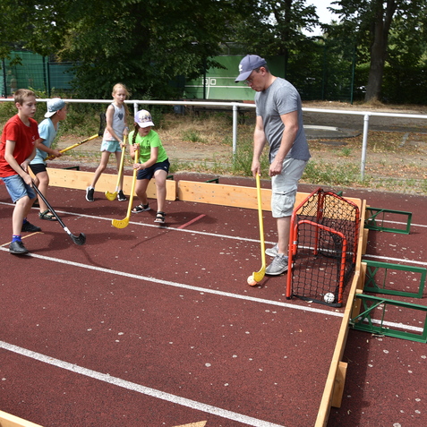 Sechs Kinder und ein Erwachsener spielen im Freien mit Hockeyschl&auml;gern auf einem roten Sportplatz.