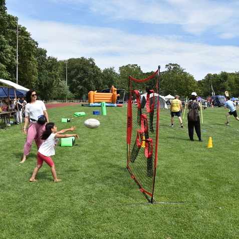 Kinder spielen Rugby auf einem gr&uuml;nen Feld, w&auml;hrend Erwachsene an einem Veranstaltungstag im Hintergrund aktiv sind.