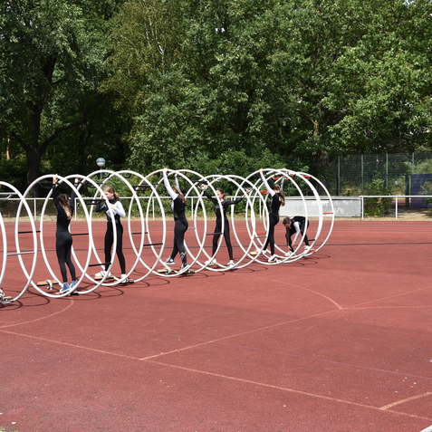 Sechs Kinder &uuml;ben mit gro&szlig;en Kunststoffreifen auf einem roten Sportplatz, umgeben von B&auml;umen.