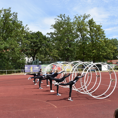 Sechs Turnerinnen f&uuml;hren synchron eine &Uuml;bung an gro&szlig;en wei&szlig;en Reifen auf einem Sportplatz aus.