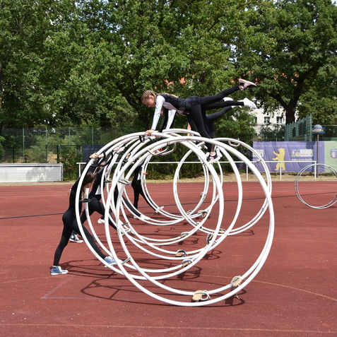 Zwei Artisten trainieren auf einem Sportplatz mit gro&szlig;en, wei&szlig;en Hula-Hoops; Zuschauer beobachten das Geschehen.