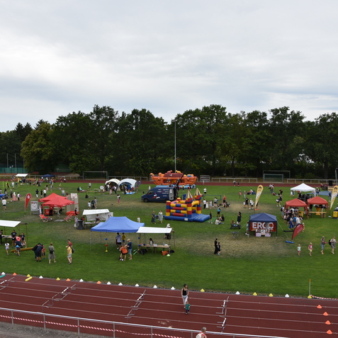Bunt dekorierte St&auml;nde und Attraktionen auf einem Sportplatz, viele Menschen genie&szlig;en ein Fest im Freien.