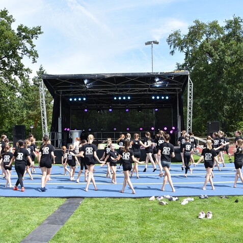 Gruppen von Tänzern in schwarzen Shirts proben auf einer blauen Fläche vor einer Bühne mit bunten Lichtern im Freien.